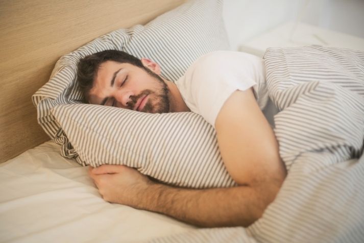 Homme aux cheveux bruns dormant paisiblement dans un lit blanc, illustrant l'importance du sommeil pour la productivité au travail.